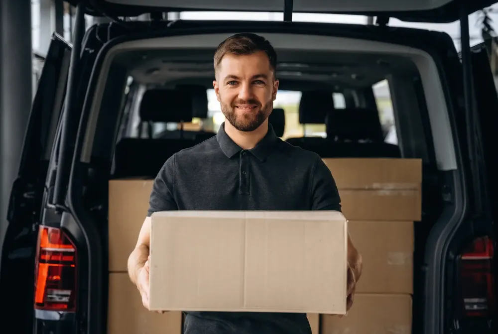 Kurier Bote l&auml;chelt in die Kamera und h&auml;lt ein Paket in der Hand. Hinter ihm steht ein Volkswagen Transporter als Firmen-Leasing, Gewerbeleasing Fahrzeug Kurierdienst